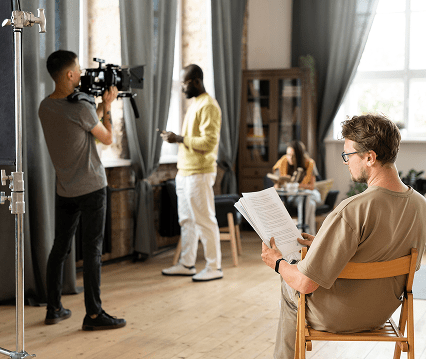 A film crew on set, with a cameraman filming a standing man, while another man sits reading a script