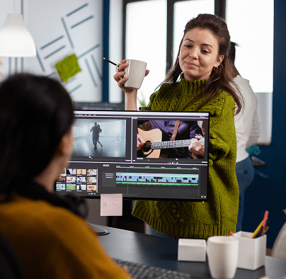 A woman in a green sweater holds a mug, looking at a coworker editing video on a desktop